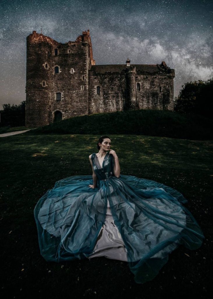 Beautiful bride in blue wedding dress sitting in front of a Castle at night with dark stormy skies